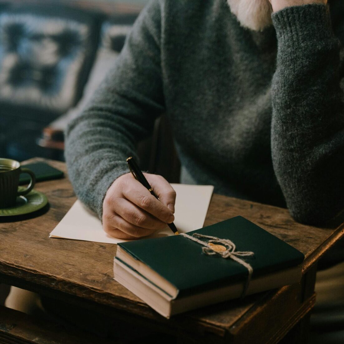 A person writes at a wooden table adorned with green decor in a cozy indoor setting.